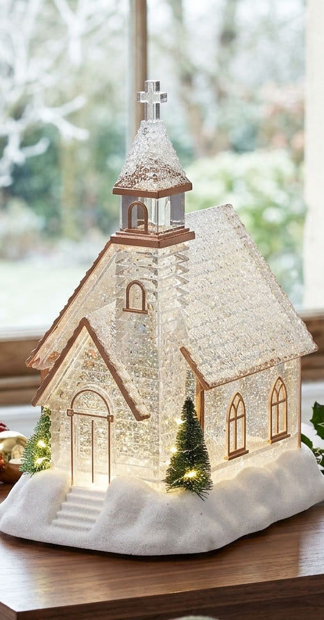 Decorative model of a church snow globe with lights on a table in front of a window with snow outside.