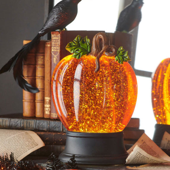 Decorative pumpkin-shaped snow globe with glowing orange light on a table with books and birds.