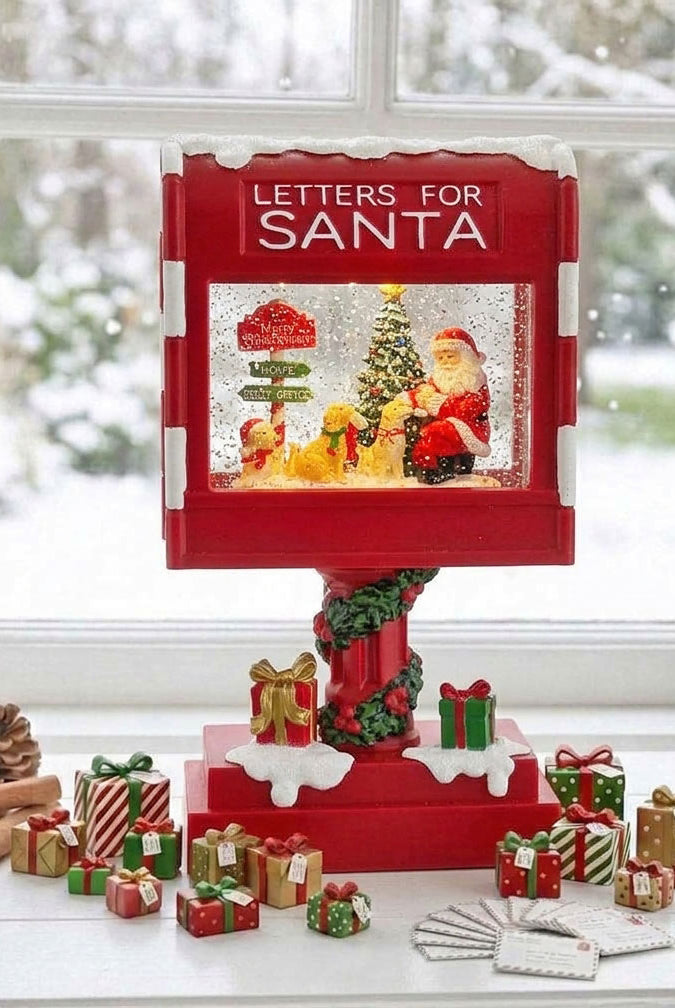 A red 'Letters for Santa' snow globe display stands on a table by a window, surrounded by small Christmas gift boxes, pinecones, cinnamon sticks, and a few folded cards. The display features Santa, a decorated tree, and falling snow inside a snow globe, with the words 'LETTERS FOR SANTA' printed on the top panel.