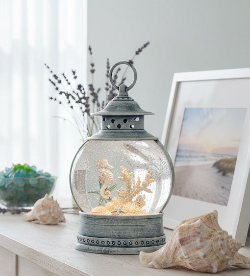 A decorative lantern-style glass globe with a metal top and base, filled with sand, seashells, coral-like pieces, and a small seahorse figure, displayed on a tabletop with seashells and a framed beach photo in a softly lit room.