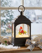 A black lantern-style holiday snow globe sits on a wooden windowsill with a snowy outdoor scene visible behind it. The lantern has a warm glowing interior with a small festive scene featuring red and green holiday decorations, white faux snow, and gold accents.