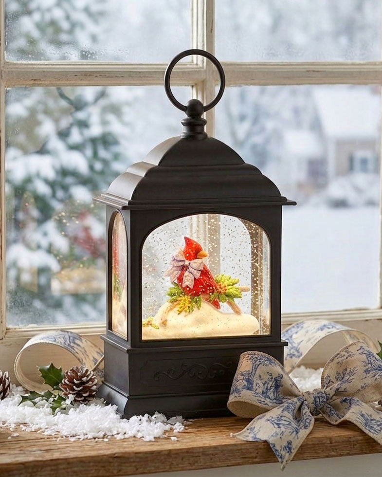 A black lantern-style holiday snow globe sits on a wooden windowsill with a snowy outdoor scene visible behind it. The lantern has a warm glowing interior with a small festive scene featuring red and green holiday decorations, white faux snow, and gold accents.