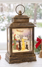 A decorative bronze lantern with a snow globe-style Christmas scene inside, featuring Santa Claus, a Christmas tree, birds, and dogs under falling snow, displayed on a windowsill with a blurred outdoor winter background.