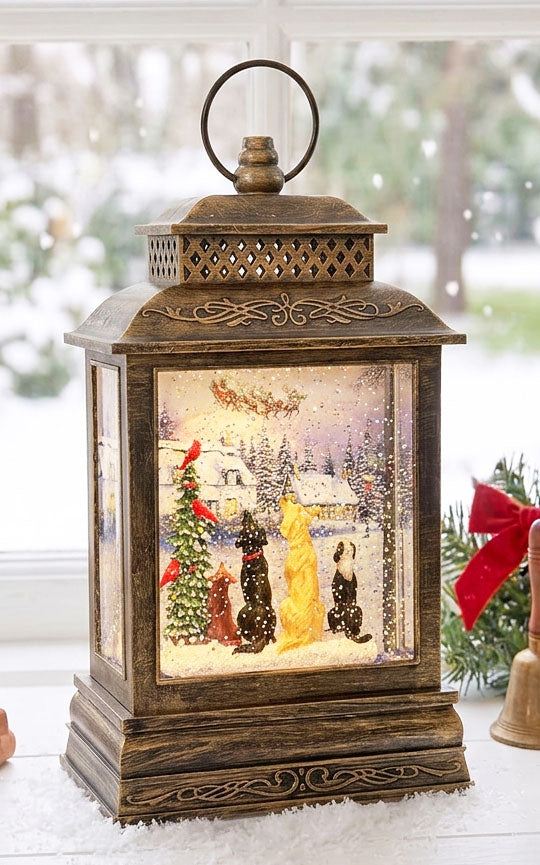 A decorative bronze lantern with a snow globe-style Christmas scene inside, featuring Santa Claus, a Christmas tree, birds, and dogs under falling snow, displayed on a windowsill with a blurred outdoor winter background.