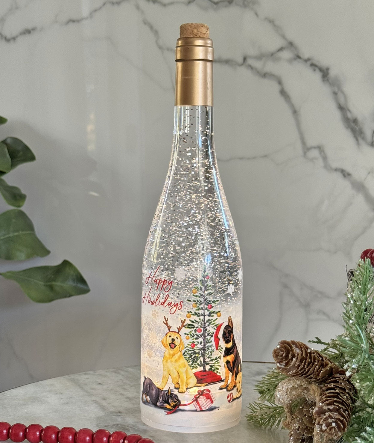 A clear glass bottle with a cork stopper, filled with glittering snow-like particles and decorated with a Christmas scene featuring two dogs, a tree, and the words 'Happy Holidays'. The bottle is displayed on a marble surface with holiday greenery and red beads in the background.