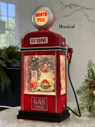 A red Christmas-themed miniature gas pump figurine with a glowing 'NORTH POLE' sign on top, a snow globe display inside, and the words 'ELF SERVE' and 'GAS STATION' on the front. The product is shown on a marble surface with holiday greenery in the background.