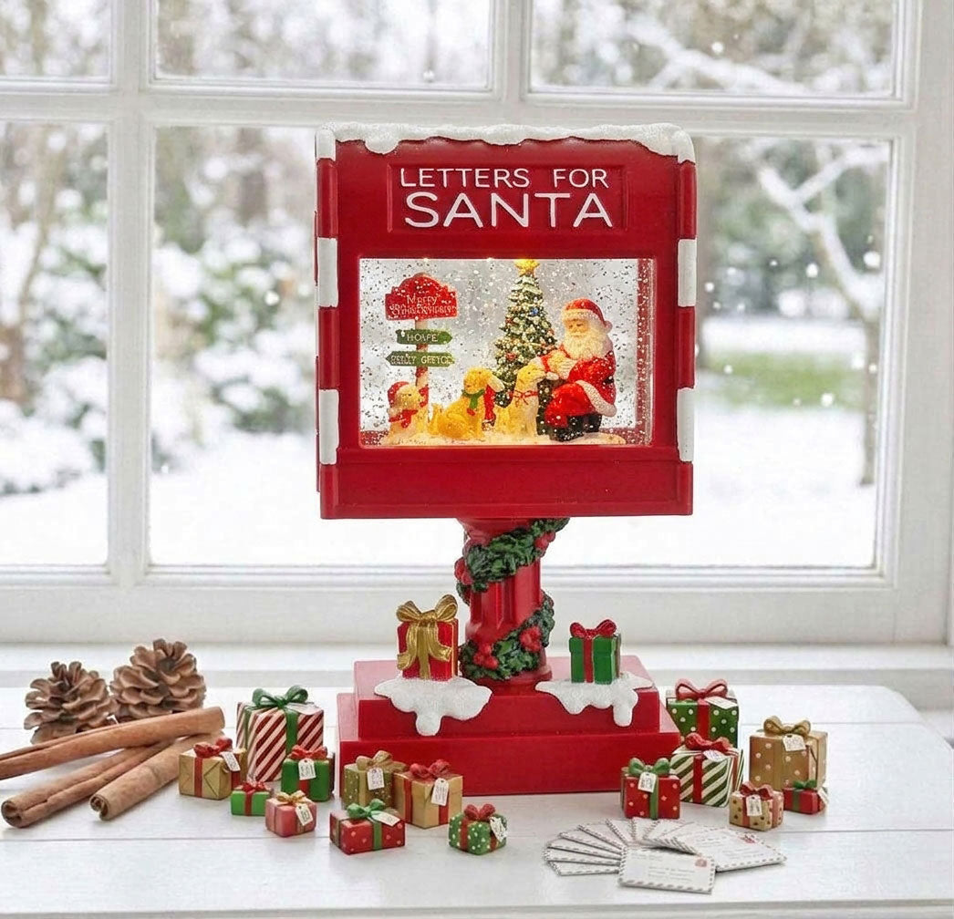 A red 'Letters for Santa' snow globe display stands on a table by a window, surrounded by small Christmas gift boxes, pinecones, cinnamon sticks, and a few folded cards. The display features Santa, a decorated tree, and falling snow inside a snow globe, with the words 'LETTERS FOR SANTA' printed on the top panel.